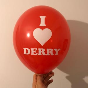 Könnte beinhalten: Ein roter Ballon mit dem weißen Text "I ♥ DERRY". Das Herzsymbol ist weiß. Der Ballon wird von einer Hand gehalten.