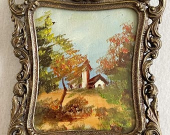 May include: A small rectangular oil painting featuring a landscape with trees and a building, enclosed in an ornate, heavy, antique-style brass frame with intricate scrollwork and floral relief patterns along the edges and corners.