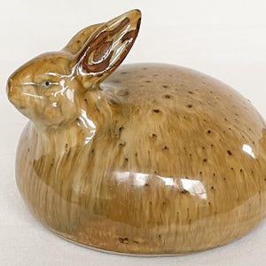 May include: A ceramic rabbit figurine in shades of brown with a glossy finish. The rabbit is in a seated position with its ears perked up. The figurine has a speckled pattern and is a decorative item.