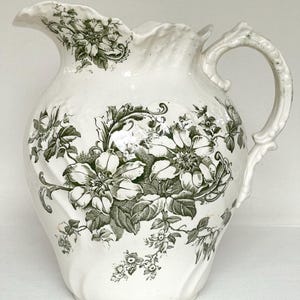 以下が含まれることがあります： クレマチスの花と葉が描かれたダークグリーンの花柄模様のクリーム色の陶器製ピッチャー。この容器には、波打つような縁、湾曲したハンドル、そして陶器の表面に成形された渦巻き状のテクスチャーが施されています。