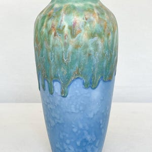 May include: A tall ceramic vase with a two-tone design. The top section has a dripped glaze in green, teal, and brown. The bottom half is a solid light blue with a speckled texture. The vase has a rounded shoulder and a narrow neck.