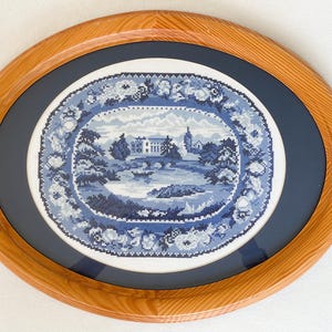 May include: An oval-shaped cross-stitch embroidery featuring a blue and white landscape scene with a building, trees, and a body of water, surrounded by a floral border. The piece is encased in a polished light brown wooden frame with a dark blue mat.