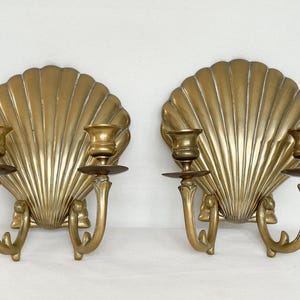 May include: A pair of antique brass wall sconces. Each sconce has a large, fan-shaped shell design with two candle holders. The sconces have a warm, golden hue and are designed for wall mounting. The candle holders are designed to hold standard-sized candles.