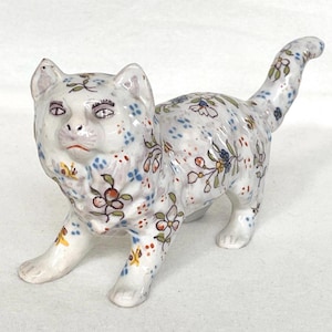 May include: A ceramic cat figurine with a white base, adorned with a floral pattern in blue, red, yellow, and green. The cat is in a walking pose with its tail raised. The figurine has detailed facial features.