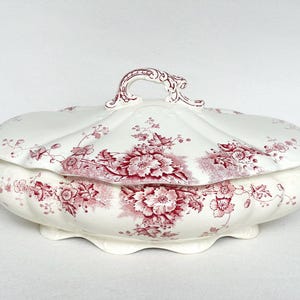 May include: A white ceramic serving dish with a lid, decorated with a red floral pattern. The dish has a scalloped edge and decorative handles. The lid features a handle. A classic piece for serving food.