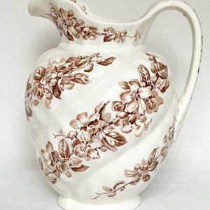 May include: A cream-coloured ceramic pitcher featuring a brown floral pattern of blossoms and leaves. The vessel has a curved handle, a wide spout, and a moulded, swirling ribbed body design that follows the diagonal orientation of the floral print.