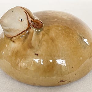 May include: A ceramic sheep ornament with a light brown body and a cream-coloured head. The sheep has a small, dark eye and brown accents around the ears and face. The ornament is glazed and has a glossy finish.