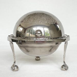 May include: A silver-colored, domed serving dish with ornate floral detailing on the lid. The dish has a round base supported by three decorative legs with ball feet. The lid has a small, round handle.