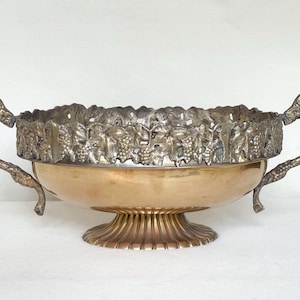 May include: A two-handled bowl featuring a smooth, polished copper-toned base and a fluted pedestal foot. The upper rim is adorned with a silver-toned metal band displaying a detailed, raised relief pattern of grape clusters and vine leaves.