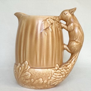 May include: A light brown ceramic pitcher featuring a vertical ribbed body and a sculpted handle shaped like a squirrel climbing a branch. The base is decorated with embossed oak leaves and acorns, and the entire piece has a glossy finish.