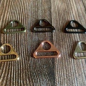 Op de afbeelding: Een verzameling van zes metalen D-ringen in verschillende afwerkingen: olijfgroen, donkergrijs, zwart, goud, koper en messing. Elke ring heeft een driehoekige vorm met een cirkelvormige opening aan de bovenkant en een rechthoekige opening aan de onderkant. De ringen worden op een houten oppervlak weergegeven.
