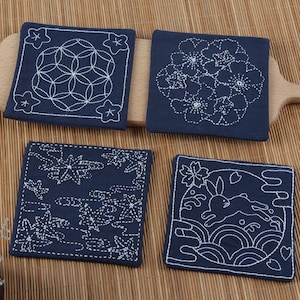 Könnte beinhalten: Vier marineblaue quadratische Untersetzer mit weißen Stickmustern. Jedes Design zeigt ein anderes Muster: geometrisch, floral, Ahornblätter und ein Hase. Diese Untersetzer schützen Oberflächen vor Flecken.