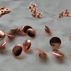 Puede incluir: Una colecci&oacute;n de botones y remaches de metal de color cobre. Los botones son redondos y tienen un centro elevado. Los remaches tienen una cabeza plana y un extremo puntiagudo.