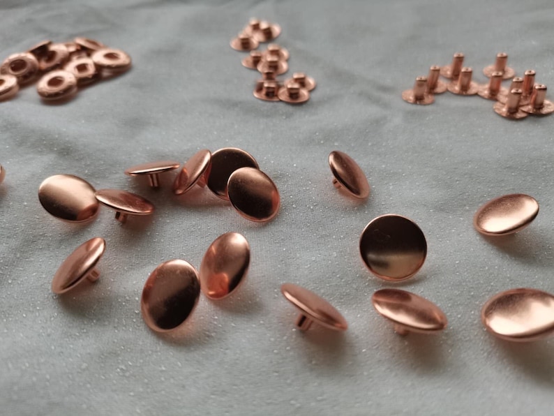 Puede incluir: Una colecci&oacute;n de botones y remaches de metal de color cobre. Los botones son redondos y tienen un centro elevado. Los remaches tienen una cabeza plana y un extremo puntiagudo.