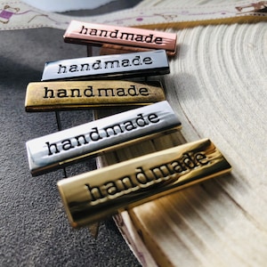 Puede incluir: Cinco etiquetas metálicas con la palabra "hecho a mano" grabada en ellas. Las etiquetas son de diferentes colores: oro rosa, plata, oro y dos tonos de latón.