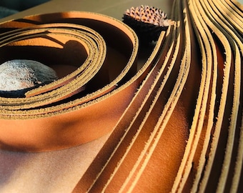 May include: A close-up of a stack of brown leather strips. The strips are arranged in a fan shape, with the edges of the strips visible. The leather is smooth and has a slight sheen.