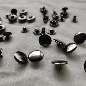 Puede incluir: Una colecci&oacute;n de botones a presi&oacute;n de metal negro, algunos con un centro elevado, esparcidos sobre una superficie de tela blanca.