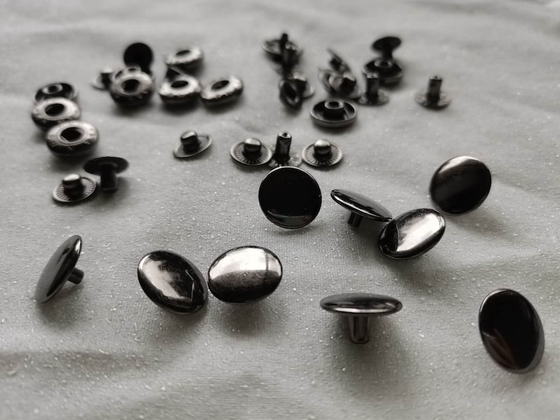 Puede incluir: Una colecci&oacute;n de botones a presi&oacute;n de metal negro, algunos con un centro elevado, esparcidos sobre una superficie de tela blanca.