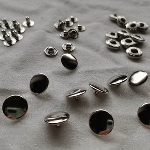Puede incluir: Una colecci&oacute;n de botones y broches de metal plateado. Los broches son redondos y tienen un agujero en el centro. Los botones son redondos y tienen una parte superior plana.