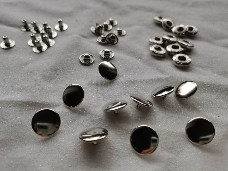 Puede incluir: Una colecci&oacute;n de botones y broches de metal plateado. Los broches son redondos y tienen un agujero en el centro. Los botones son redondos y tienen una parte superior plana.