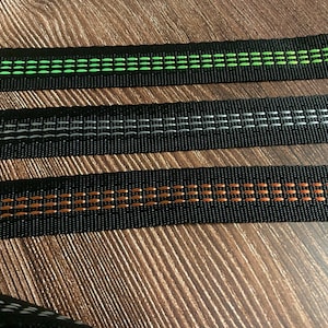 May include: Close-up of three black nylon straps with decorative stitching. The top strap has green stitching, the middle strap has white stitching, and the bottom strap has orange stitching. The straps are lying on a wooden surface.