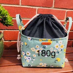 K&ouml;nnte beinhalten: Eine gr&uuml;ne Kordeltasche mit wei&szlig;em Blumendruck und schwarzem Futter. Die Tasche hat zwei Henkel und ein braunes Leder-Etikett mit der Aufschrift "Hand Made". Die Tasche wiegt 180 Gramm.