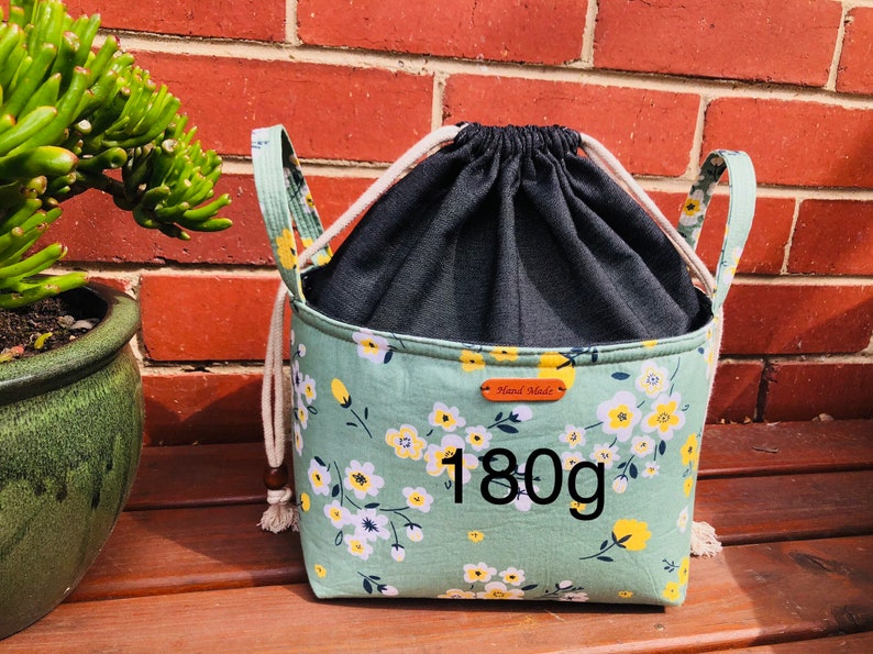 K&ouml;nnte beinhalten: Eine gr&uuml;ne Kordeltasche mit wei&szlig;em Blumendruck und schwarzem Futter. Die Tasche hat zwei Henkel und ein braunes Leder-Etikett mit der Aufschrift "Hand Made". Die Tasche wiegt 180 Gramm.