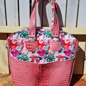 K&ouml;nnte beinhalten: Eine rosafarbene Tasche mit Kroko-Pr&auml;gung und einem Blumendruck mit rosa und grauen V&ouml;geln. Die Tasche hat zwei Henkel oben und einen abnehmbaren Schultergurt.