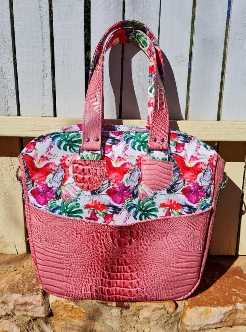 K&ouml;nnte beinhalten: Eine rosafarbene Tasche mit Kroko-Pr&auml;gung und einem Blumendruck mit rosa und grauen V&ouml;geln. Die Tasche hat zwei Henkel oben und einen abnehmbaren Schultergurt.