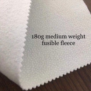 K&ouml;nnte beinhalten: Wei&szlig;er, schmelzbarer Fleece-Stoff mit gepunkteter Textur. Der Stoff ist mit "180g mittelschwerer schmelzbarer Fleece" beschriftet.