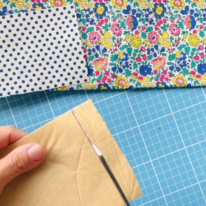 Puede incluir: Primer plano de una mano sosteniendo un sobre de papel marr&oacute;n con una herramienta blanca que lo corta. El sobre est&aacute; sobre una estera de corte azul con cuadr&iacute;cula.