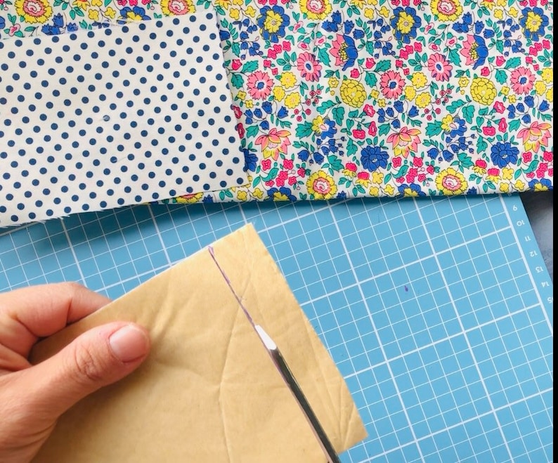 Puede incluir: Primer plano de una mano sosteniendo un sobre de papel marr&oacute;n con una herramienta blanca que lo corta. El sobre est&aacute; sobre una estera de corte azul con cuadr&iacute;cula.