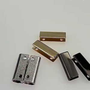 May include: A selection of metal shoe aglets in different finishes. The aglets are rectangular with rounded edges and small holes for attaching to shoelaces. Finishes include silver, gold, and black.
