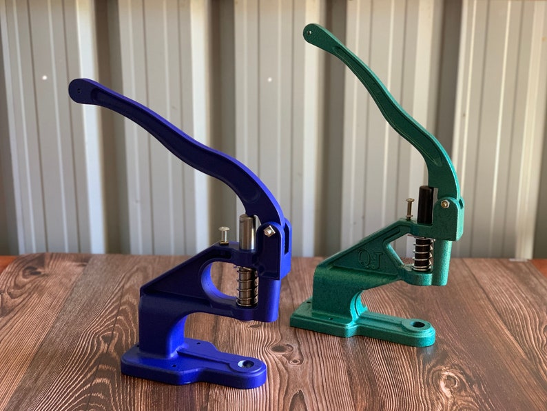 Op de afbeelding: Twee metalen knoopsteker, een blauwe en een groene, met een hendel en een veermechanisme. De gereedschappen liggen op een houten oppervlak.