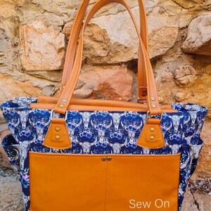 K&ouml;nnte beinhalten: Eine gro&szlig;e braune Ledertasche mit einem blauen und wei&szlig;en Blumendruck mit wei&szlig;en Totenk&ouml;pfen. Die Tasche hat eine gro&szlig;e Fronttasche und zwei obere Griffe. Der Text "Sew On Flinders" ist auf der Fronttasche aufgedruckt.