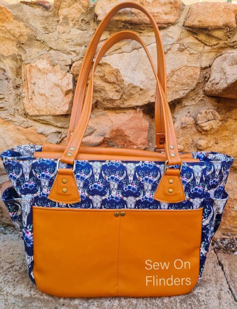 K&ouml;nnte beinhalten: Eine gro&szlig;e braune Ledertasche mit einem blauen und wei&szlig;en Blumendruck mit wei&szlig;en Totenk&ouml;pfen. Die Tasche hat eine gro&szlig;e Fronttasche und zwei obere Griffe. Der Text "Sew On Flinders" ist auf der Fronttasche aufgedruckt.