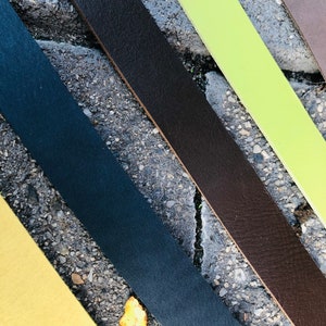K&ouml;nnte beinhalten: F&uuml;nf Lederriemen in verschiedenen Farben: Braun, Gr&uuml;n, Schwarz, Gelb und Dunkelblau. Die Riemen sind diagonal auf einer grauen Betonfl&auml;che angeordnet.