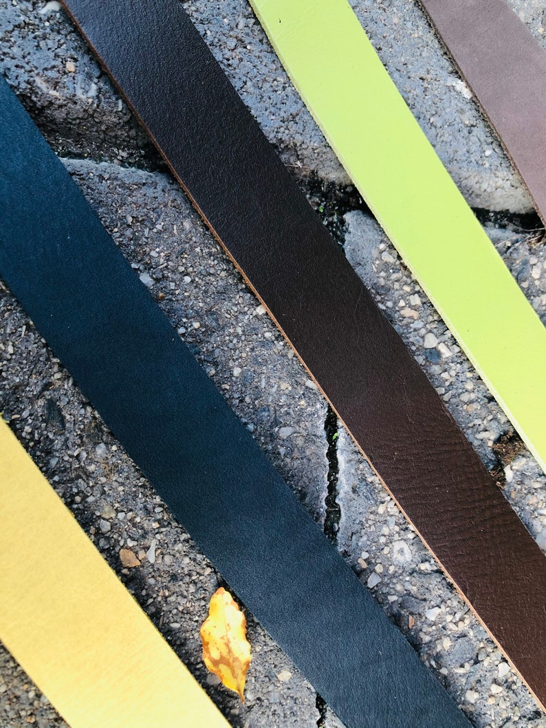 K&ouml;nnte beinhalten: F&uuml;nf Lederriemen in verschiedenen Farben: Braun, Gr&uuml;n, Schwarz, Gelb und Dunkelblau. Die Riemen sind diagonal auf einer grauen Betonfl&auml;che angeordnet.