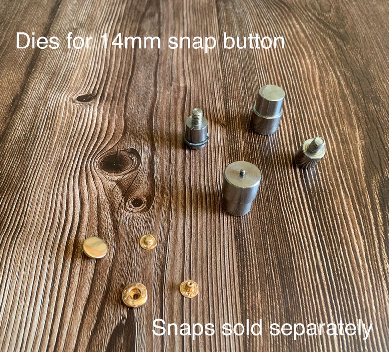 Op de afbeelding: Set metalen matrijzen voor het plaatsen van 14 mm drukknopen. De matrijzen zijn zilverkleurig en hebben een gladde afwerking. De afbeelding toont ook vier goudkleurige drukknopen. De tekst "Dies for 14mm snap button" en "Snaps sold separately" is zichtbaar in de afbeelding.