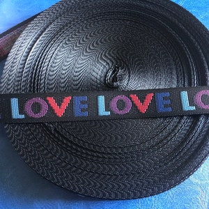 May include: Black webbing with the word "LOVE" repeated in colorful letters. The webbing is rolled up and ready to be used for crafting projects.