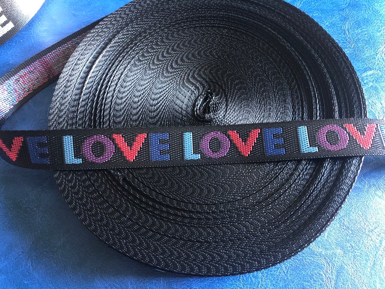 May include: Black webbing with the word "LOVE" repeated in colorful letters. The webbing is rolled up and ready to be used for crafting projects.