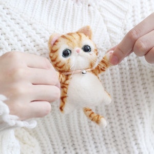 May include: A small, orange and white felted cat with blue eyes and a pink collar with a silver bell. The cat is being held by a person's hands.