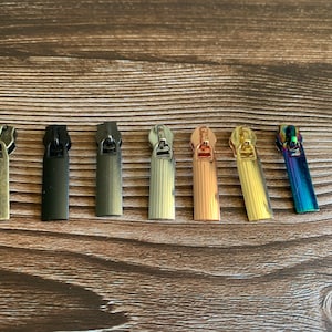 May include: A collection of seven metal zipper pulls in various colors, including antique brass, black, gunmetal, silver, rose gold, gold, and iridescent. The pulls are rectangular with a small loop at the top for attachment.
