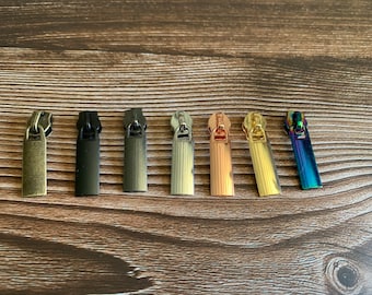 Könnte beinhalten: Eine Sammlung von sieben Metall-Reißverschluss-Anhängern in verschiedenen Farben, darunter Antik-Messing, Schwarz, Gunmetal, Silber, Roségold, Gold und schillernd. Die Anhänger sind rechteckig mit einer kleinen Schlaufe zum Befestigen.