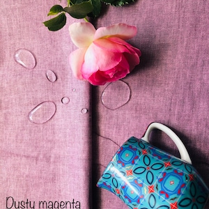 K&ouml;nnte beinhalten: Ein staubiger magentafarbener Stoff mit einer rosa Rose und Wassertropfen. Ein blauer und roter, gemusterter Keramik Krug mit einem wei&szlig;en Henkel befindet sich auf dem Stoff.