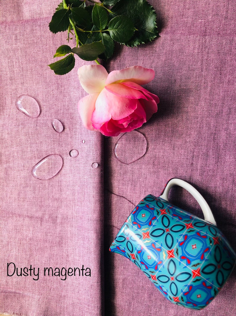 K&ouml;nnte beinhalten: Ein staubiger magentafarbener Stoff mit einer rosa Rose und Wassertropfen. Ein blauer und roter, gemusterter Keramik Krug mit einem wei&szlig;en Henkel befindet sich auf dem Stoff.