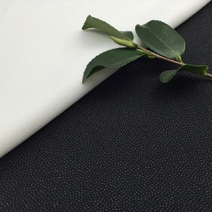 May include: A close-up of two fabric swatches, one white and one black. The white fabric is smooth and the black fabric has a textured, dotted pattern. A green leaf branch is draped over the fabric swatches.