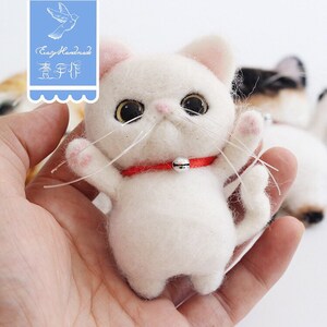 May include: A white felted cat with pink paws and a red collar with a silver bell. The cat is standing on its hind legs with its front paws raised.