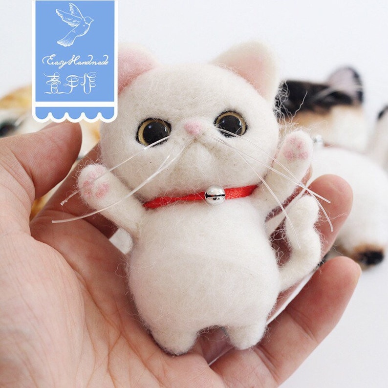 May include: A white felted cat with pink paws and a red collar with a silver bell. The cat is standing on its hind legs with its front paws raised.