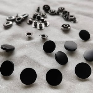 Puede incluir: Una colecci&oacute;n de botones a presi&oacute;n de metal negro, algunos con detalles plateados, sobre un fondo de tela blanca.